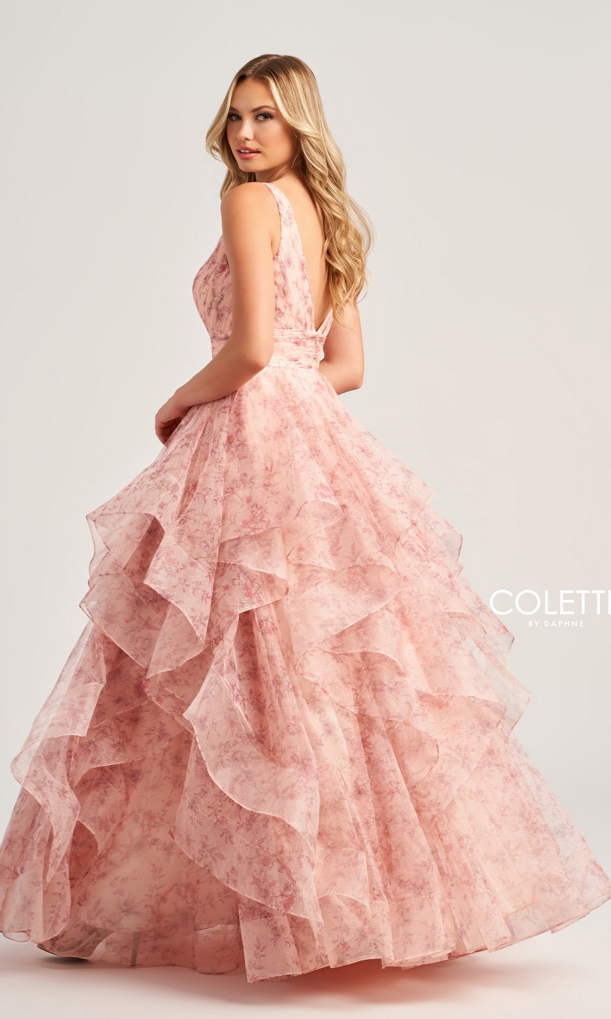 A woman poses against a plain background wearing the Colette Tiered Long Print Prom Ball Gown CL5273, a sleeveless ball gown featuring pink floral print and layered ruffles.